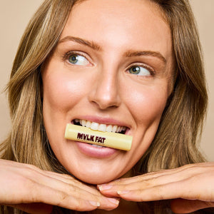 Woman holding a 'Mylk Fat' lip balm in front of her mouth against a beige background